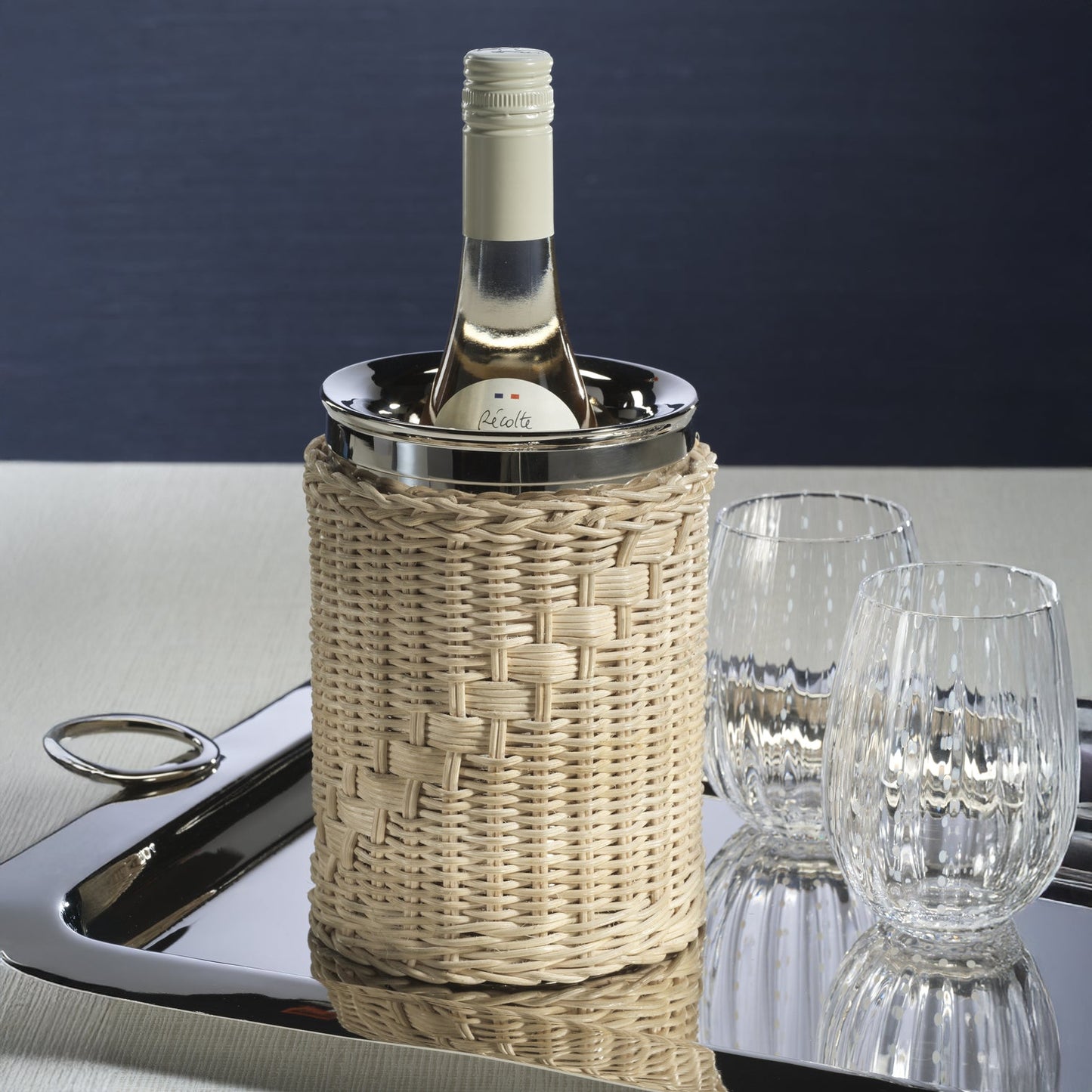 The Palmier Wine Cooler with handwoven rattan exterior and polished metal interior, holding a bottle of white wine on a tray beside glasses.