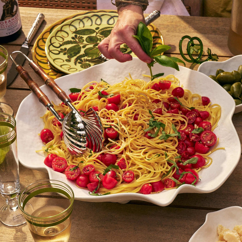 A large white scalloped serving platter filled with spaghetti, cherry tomatoes, and green herbs on a rustic wooden table, with a hand sprinkling basil over the pasta. Dining glasses, a bottle, and other tableware are visible around it.
