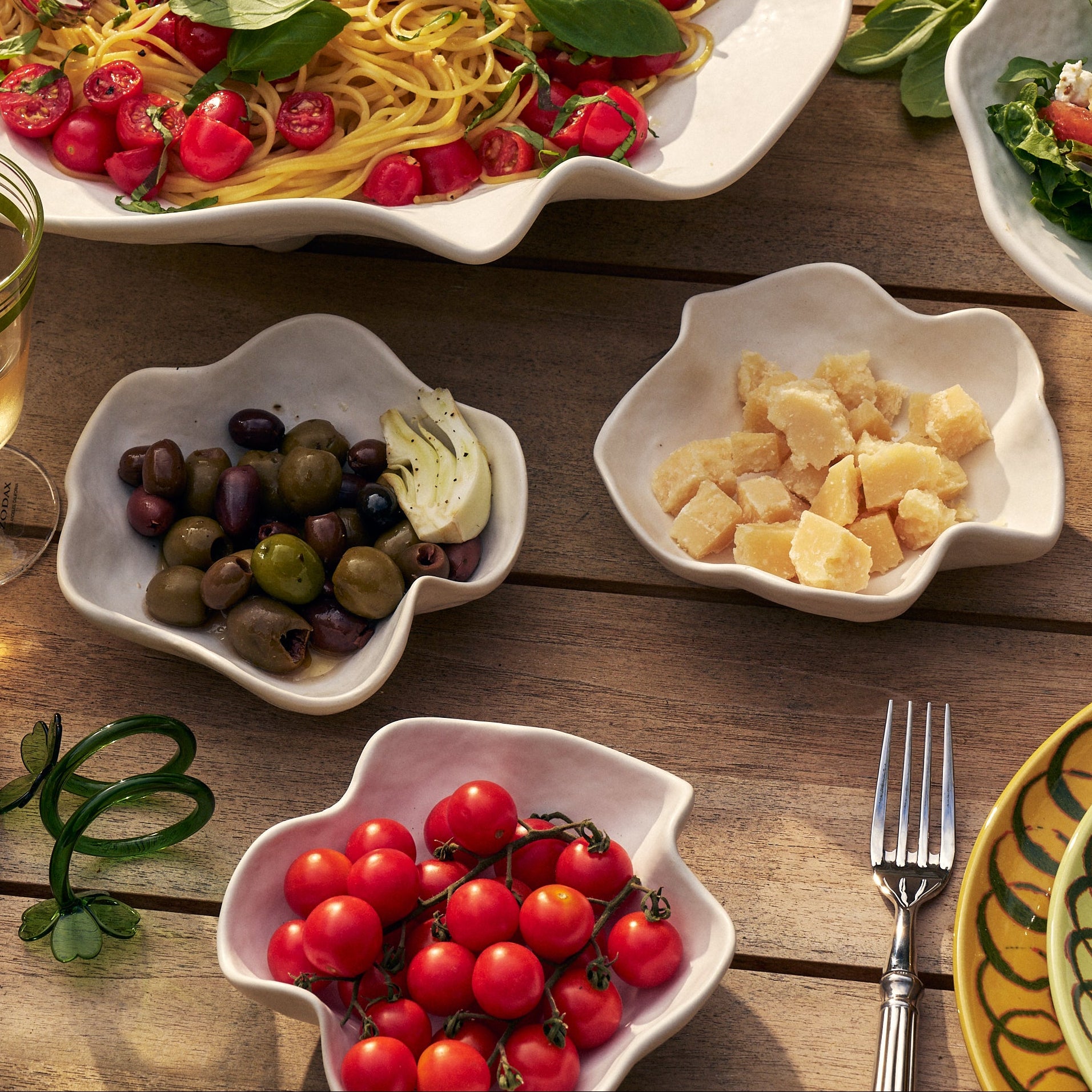 A styled outdoor dining table featuring a large white scalloped platter of spaghetti with cherry tomatoes and basil, surrounded by smaller white dishes of olives, garlic, and cherry tomatoes, alongside patterned yellow and green plates, glasses of white wine, and a wicker chair in the background.