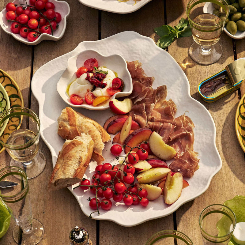 A styled dining table scene featuring a large white scalloped serving platter filled with sliced cured meat, bread, apple slices, and red berries, surrounded by glasses, plates, olives, and other dishes on a wooden table.