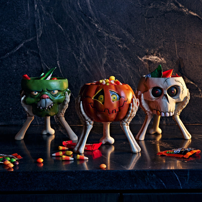 Three Halloween-themed candy bowls shaped like a green monster, a jack-o'-lantern, and a skull, filled with candy and placed on a dark countertop against a dark marble background.