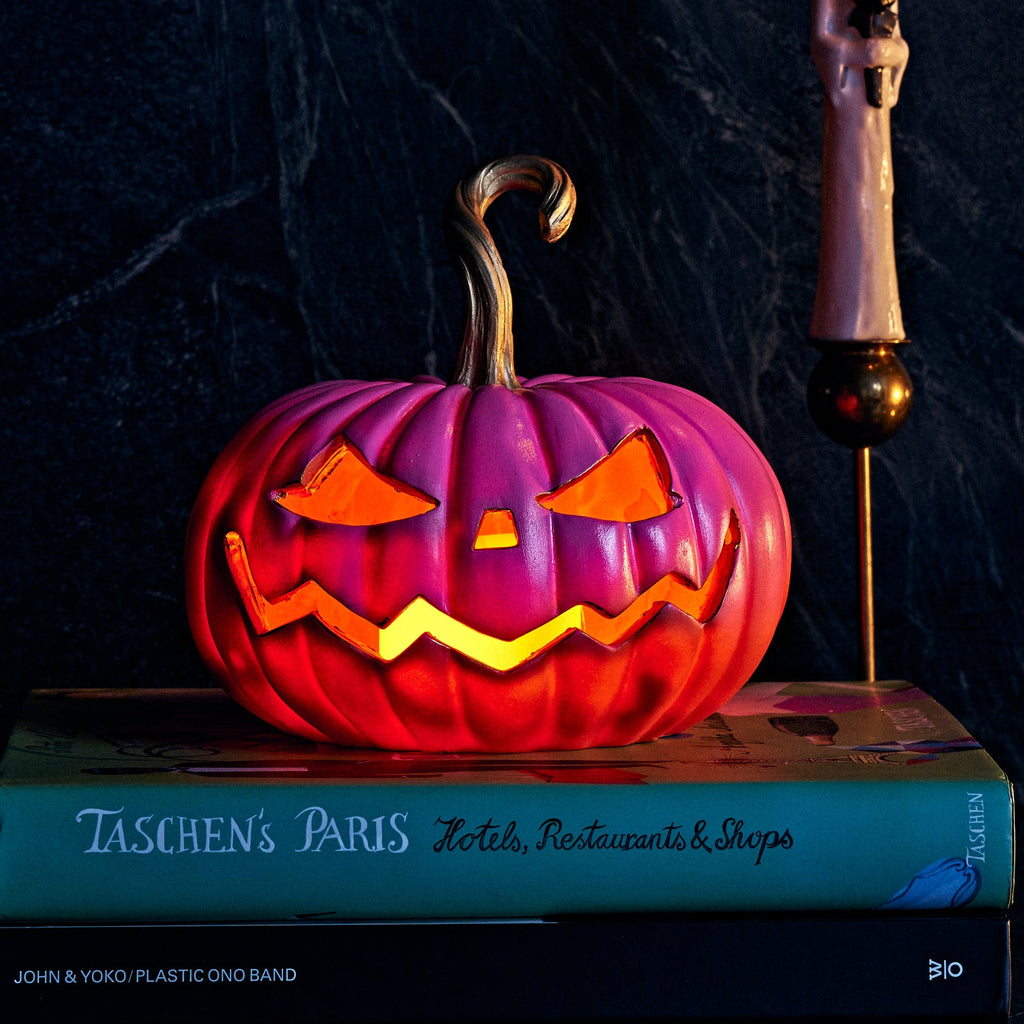 A Halloween-themed tabletop scene with a carved jack-o'-lantern glowing on a book, two lit decorative candles on stands, and a small skeleton figurine against a dark marbled background.