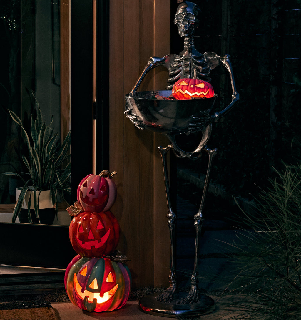 Halloween outdoor decor scene with a tall black skeleton holding a glowing jack-o'-lantern bowl, next to a stacked trio of carved pumpkins glowing with warm light on a patio at night.
