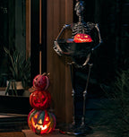 Halloween outdoor decor scene with a tall black skeleton holding a glowing jack-o'-lantern bowl, next to a stacked trio of carved pumpkins glowing with warm light on a patio at night.