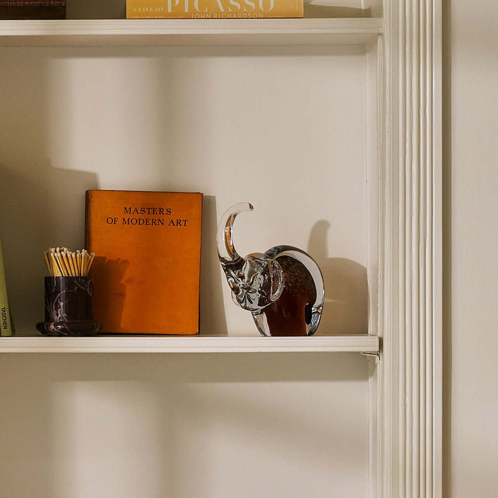 Great Elephant - Golden Amber Glass sculpture displayed on a bookshelf alongside art books.