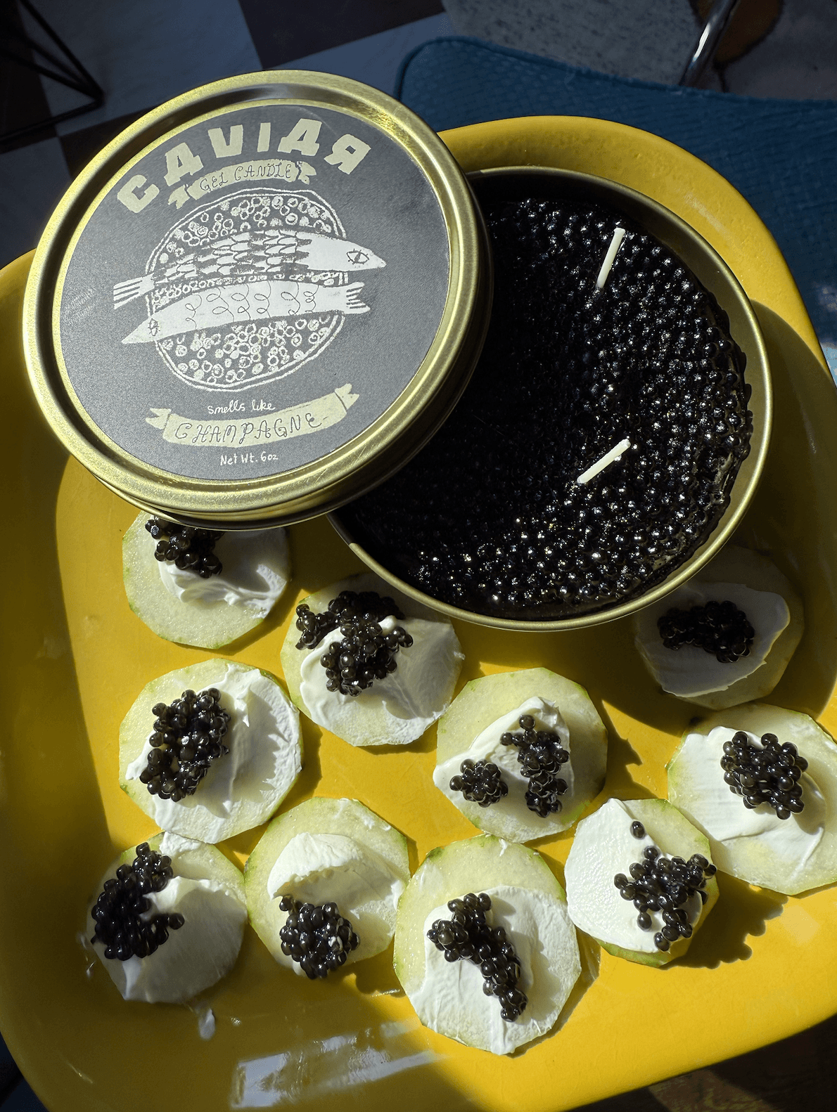 Fancy Caviar Candle in a caviar tin with gel wax candle and caviar-topped appetizers on a yellow plate.