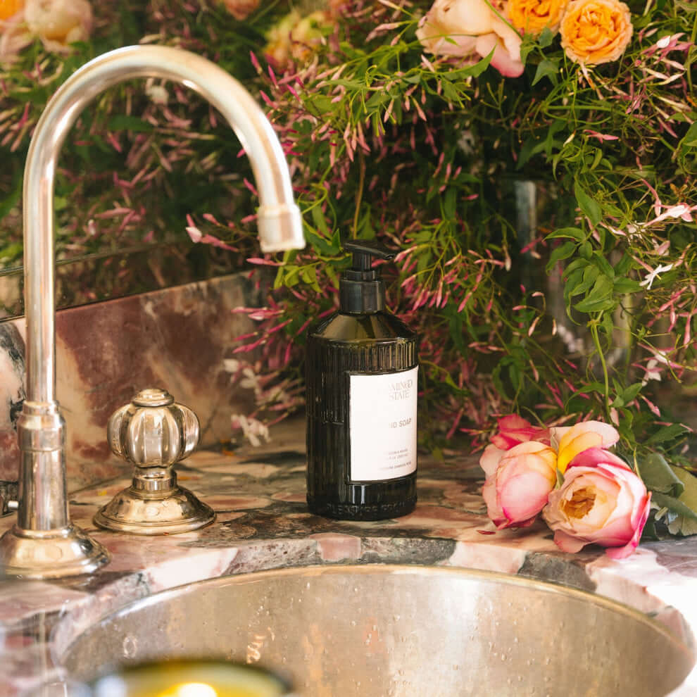 Jasmine Damask Rose Glass Hand Soap displayed beside a sink with floral arrangements in the background.