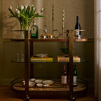 Sloane Oak Wood Oval Bar Cart styled with drinks, candle holders, and flowers in an elegant setting.