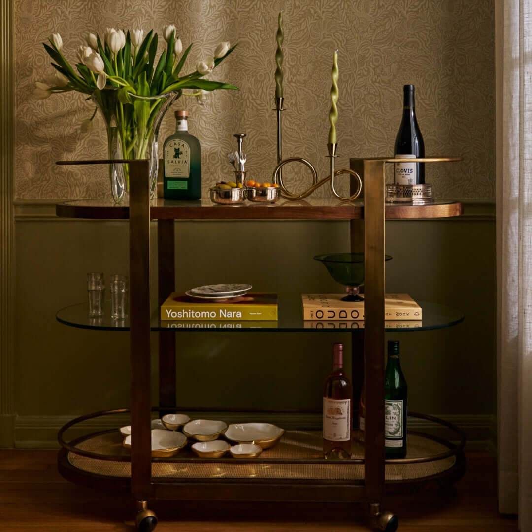 Sloane Oak Wood Oval Bar Cart styled with drinks, candle holders, and flowers in an elegant setting.