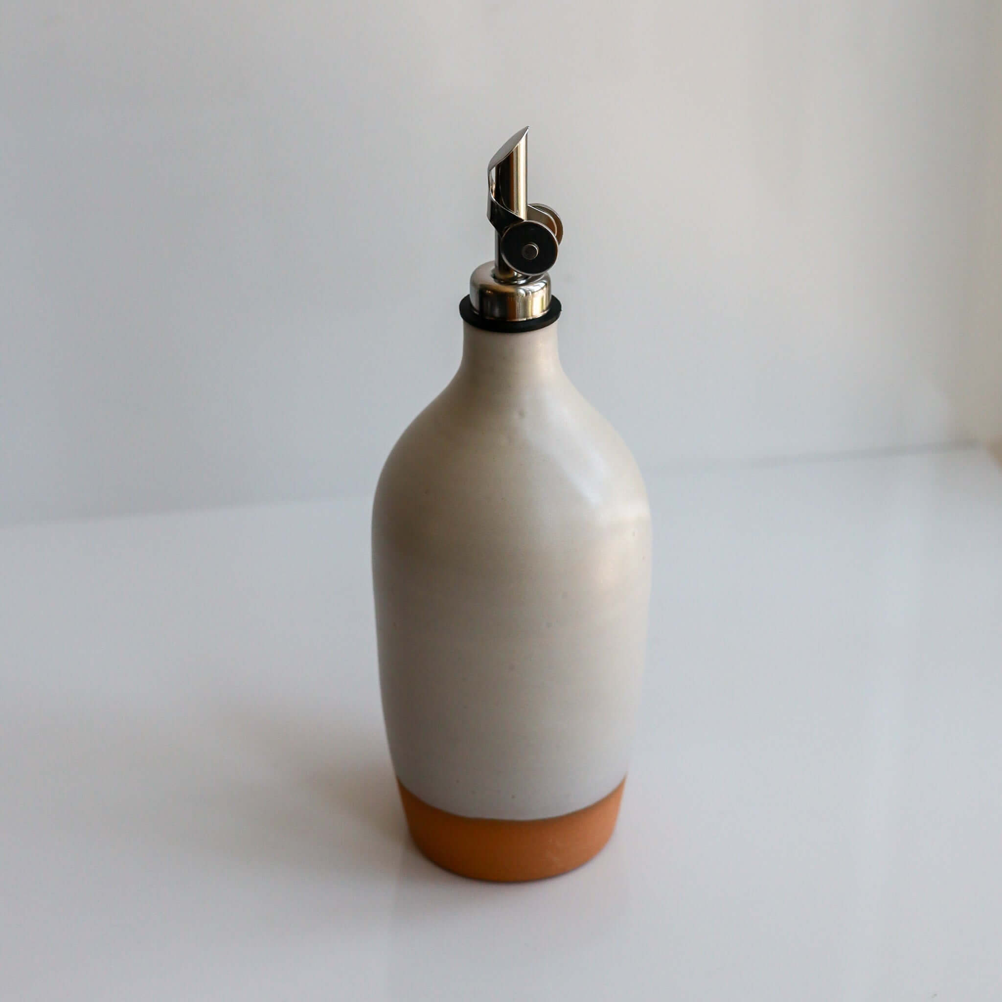 Tall Handmade Ceramic Oil Bottle in matte white with polished pourer and terracotta base on a counter.