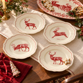 Four scalloped ceramic plates with red deer illustrations and red border detailing, arranged on a table runner with holiday decorations and cookies.
