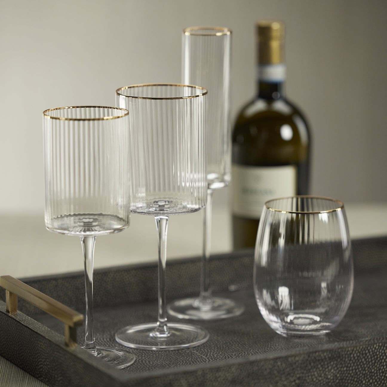 Set of clear wine glasses with gold rims on a tray, with a bottle of wine in the background.