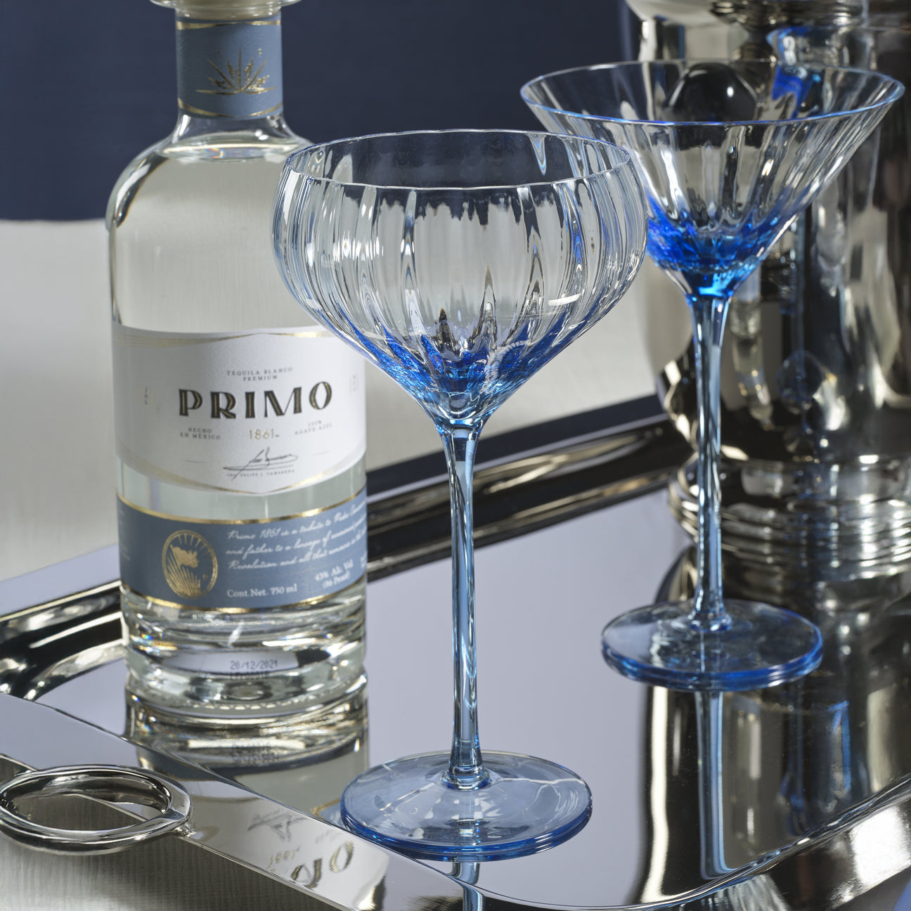 Bottle of Primo tequila with two blue-tinted cocktail glasses on a reflective surface.