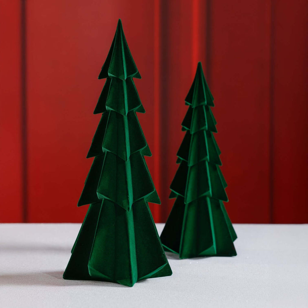 Two green paper trees on a white surface with a red curtain background
