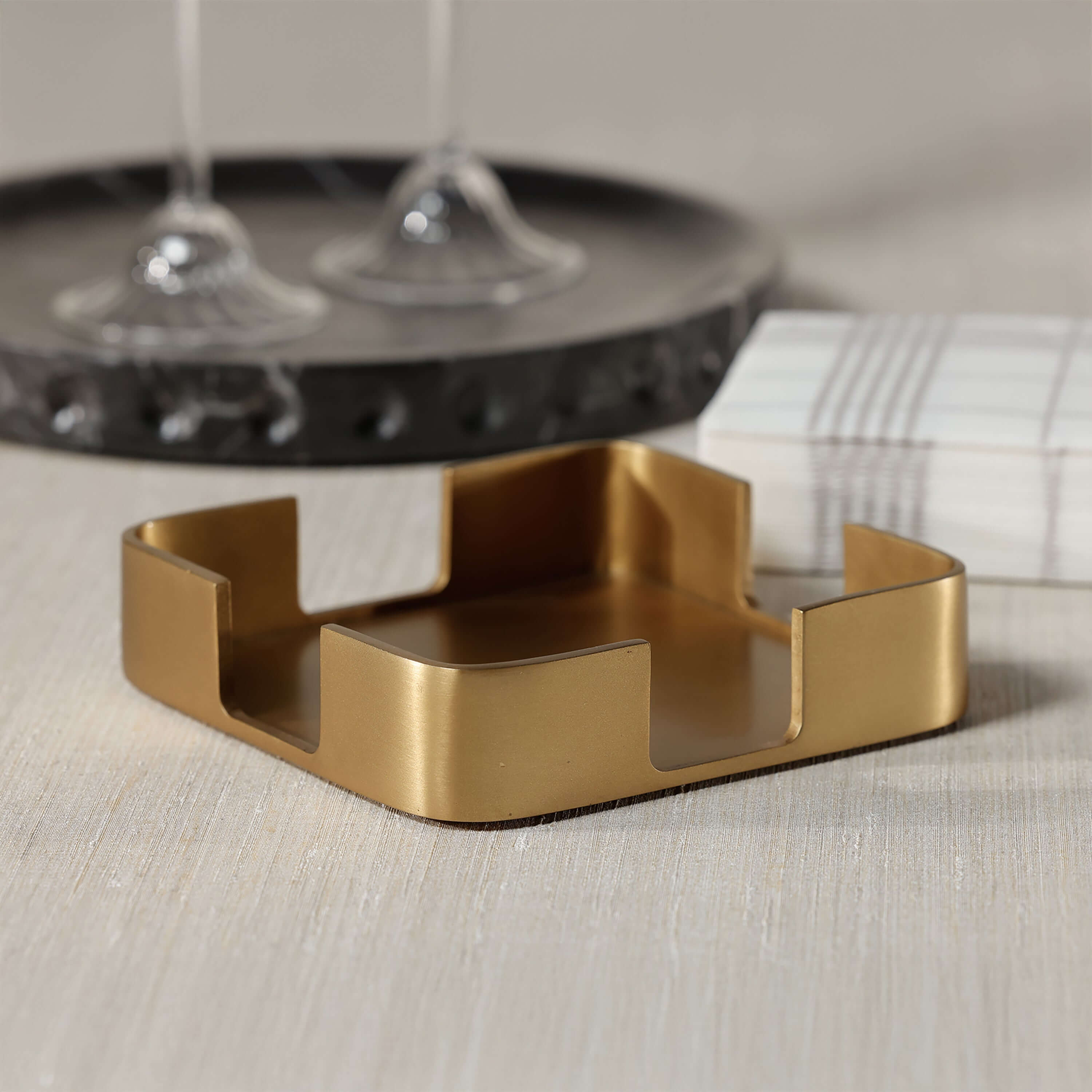 Gold Cocktail Napkin Holder with satin brushed gold finish on a tabletop next to cocktail glasses.