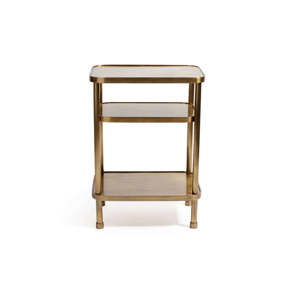 Aman Étagère, a three-tier open shelving table in warm antique brass, featuring rounded corners and cylindrical posts.