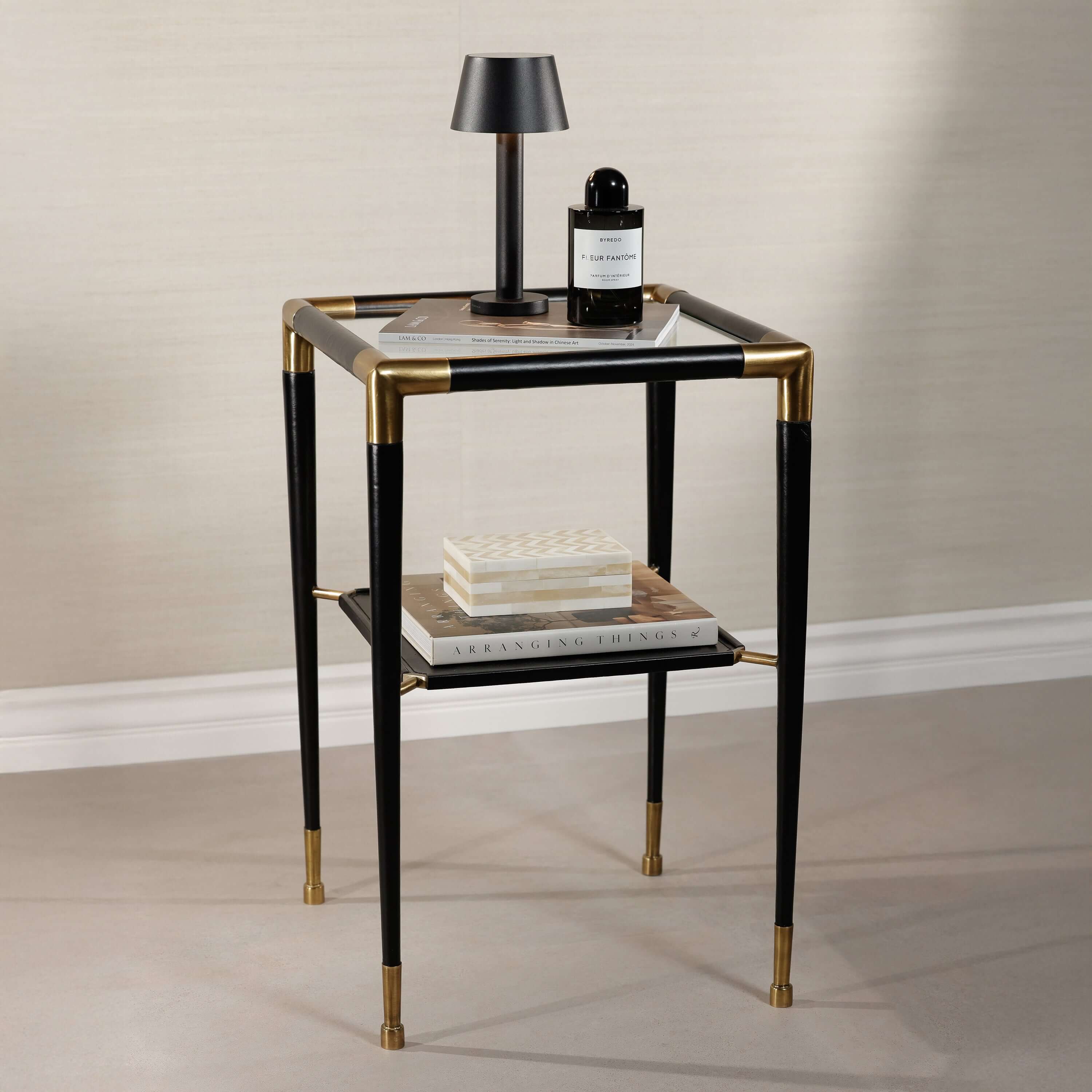 Peninsula Leather Side Table with glass top, black leather finish, and brass accents, styled with books and a lamp.