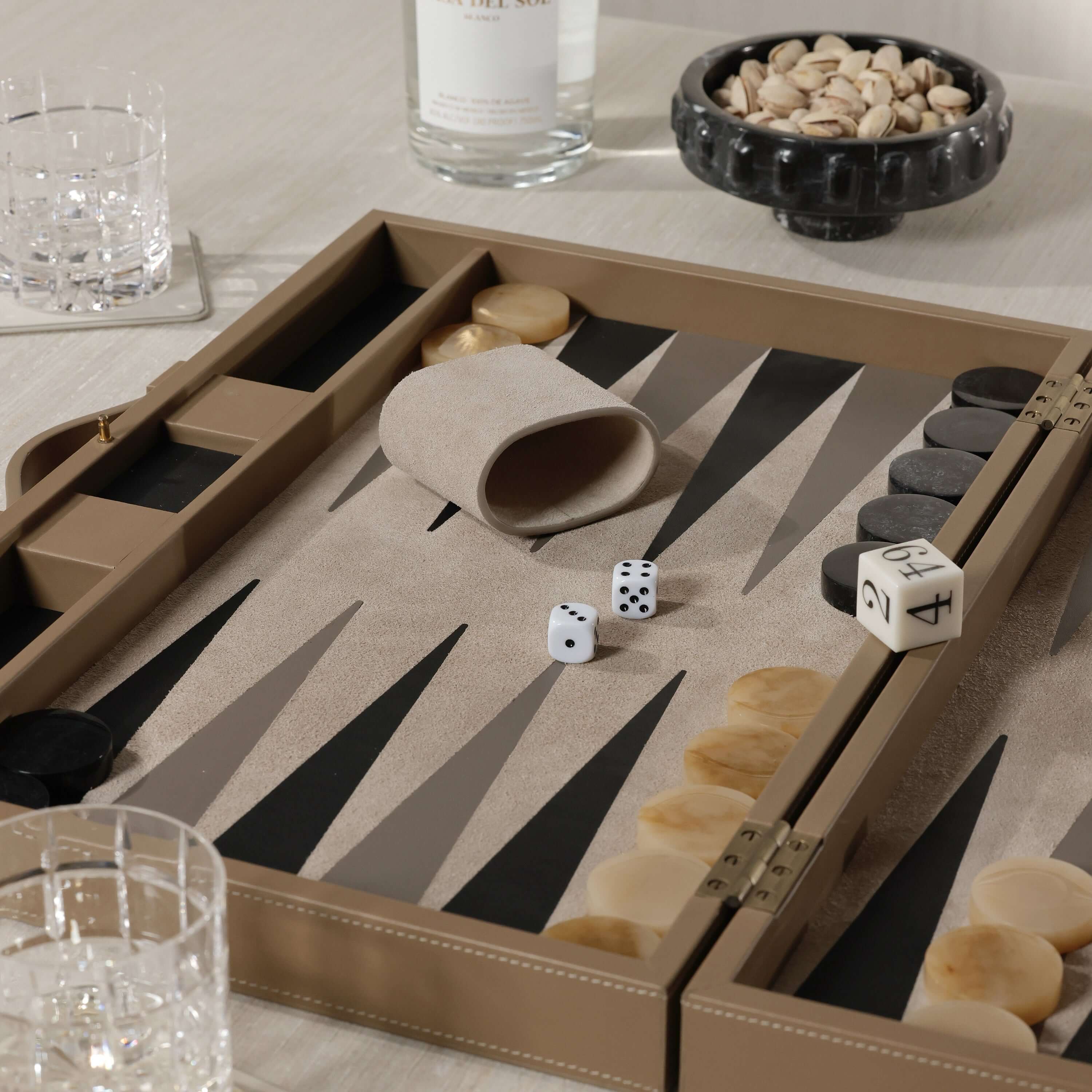 Geneva 19" Backgammon Set - Taupe Leather with dice on a smooth playing surface and elegant drinkware in the background.
