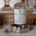 Sur Mer Shell Design Bottle Coaster holding a bottle of Flecha Azul Tequila Blanco with a clear glass next to it.