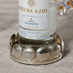Sur Mer Shell Design Bottle Coaster showcasing an elegant silver shell motif holding a tequila bottle on a textured table.