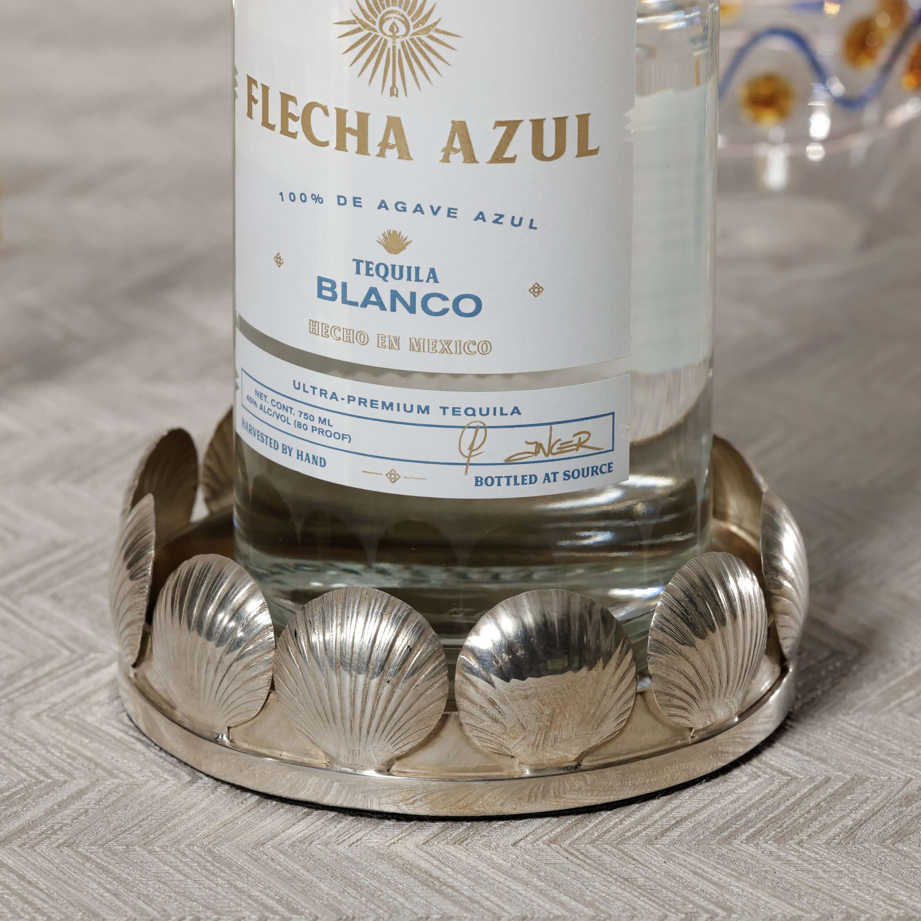 Sur Mer Shell Design Bottle Coaster showcasing an elegant silver shell motif holding a tequila bottle on a textured table.