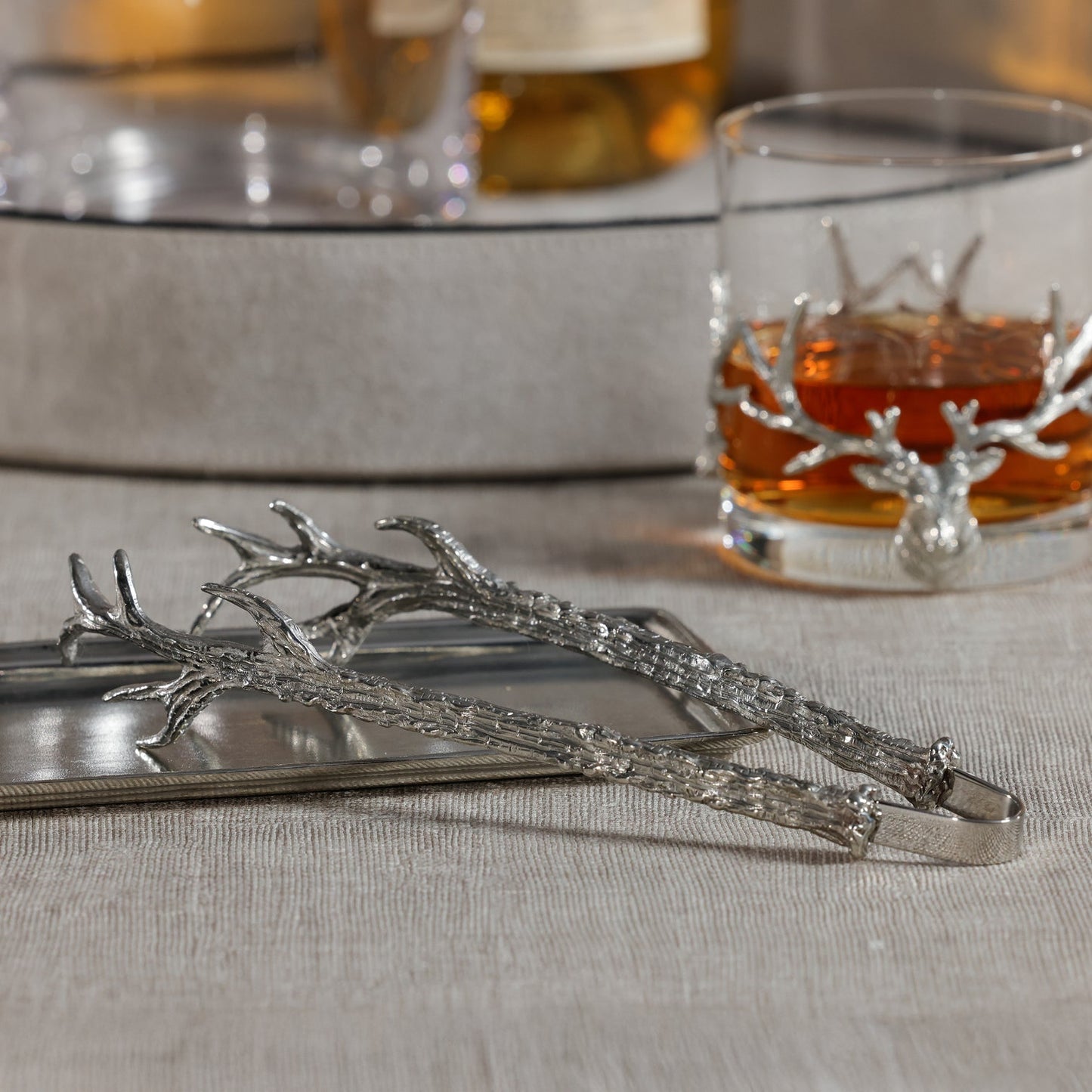 Silver branch-shaped tongs on a surface with a glass of amber liquid in the background.
